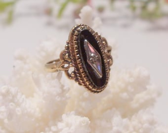 Pode incluir: Um anel de ouro com uma pedra oval preta engastada em um design de filigrana dourada. A pedra tem uma pedra menor em forma de diamante no centro.