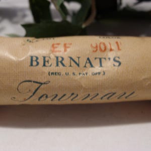 May include: Close-up of a vintage Bernat's Tournan yarn label. The label is tan with the name "BERNAT'S" in blue, and "Tournan" in a script font. The label also has the text "(REG. U.S. PAT. OFF.)" and the number "9011".