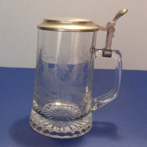 May include: A clear glass beer stein with a metal hinged lid and handle. The glass is etched with a pheasant and foliage design. The base has a decorative, scalloped edge.