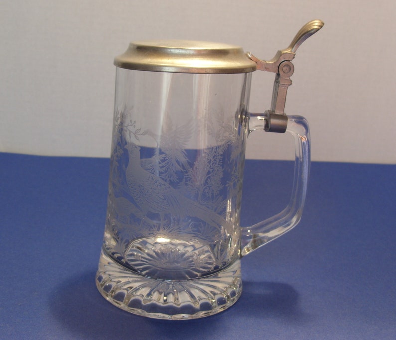 May include: A clear glass beer stein with a metal hinged lid and handle. The glass is etched with a pheasant and foliage design. The base has a decorative, scalloped edge.