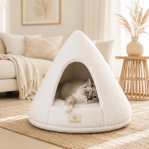 Peut inclure: Un lit pour animal de compagnie en forme de cône, de couleur crème, fabriqué en polaire texturée douce. Le lit présente une ouverture triangulaire avec un chat de couleur claire à l'intérieur. Une étiquette rectangulaire porte le texte RAPUTKI SHOP.