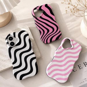 Puede incluir: Tres fundas para smartphone con bordes ondulados y patrones repetitivos llamativos. Una funda tiene ondas negras y blancas, otra ondas negras y rosas, y la tercera ondas de color rosa claro y blanco. Cada funda incluye un recorte para la lente.