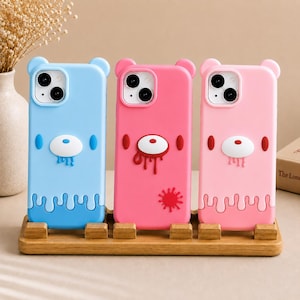 Puede incluir: Tres fundas de silicona para teléfono con orejas de oso en la parte superior. La funda de la izquierda es azul claro con un hocico blanco y gotas azules. La del medio es rosa intenso con un hocico blanco, gotas rojas y una mancha roja. La de la derecha es rosa claro con un hocico blanco y gotas rosas.