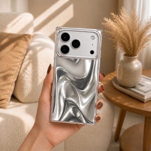 May include: A rectangular smartphone case featuring a clear frame and a back panel with a liquid-like, metallic silver wavy pattern. The upper section includes a cutout for a triple-lens camera system and a flash, maintaining a sleek, reflective finish.