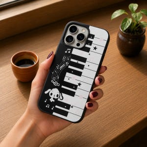 May include: A black and white smartphone case featuring a piano keyboard pattern, musical notes, small stars, and a cartoon character with long ears. The design includes a treble clef and scattered musical symbols across the keys.