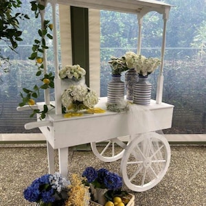 Könnte beinhalten: Ein weißer Holzwagen mit Überdachung, ausgestattet mit vier Speichenrädern und Griffen. Der Wagen ist mit weißen Blumenarrangements, blau-weiß gestreiften Vasen und einer Ranke mit gelben Zitronen geschmückt. Zwei Holzkisten mit blauen Hortensien und gelben Zitronen stehen am Boden.