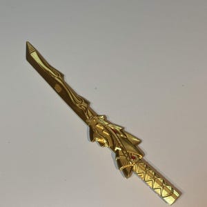 Op de afbeelding: Een goudkleurig decoratief zwaard met een puntig lemmet, een handvat met een ruitpatroon en een centraal geplaatst stootplaatgedeelte versierd met gelaagde, vleugelachtige uitsteeksels en een kleine, ingebedde rode cirkelvormige accent.