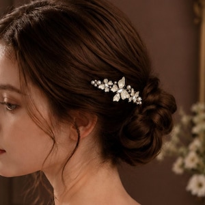 May include: A hair comb featuring a central butterfly motif adorned with white petals, small clear crystals, and round white pearls. The decorative piece is secured into a dark brown braided bun hairstyle, showcasing a symmetrical arrangement of floral-inspired elements and shimmering accents.