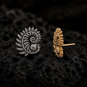 May include: Two stud earrings featuring a detailed fern frond design that curls into a spiral shape. One earring is shown from the front in a silver tone, while the other is shown from the side in a gold tone, revealing the post backing.