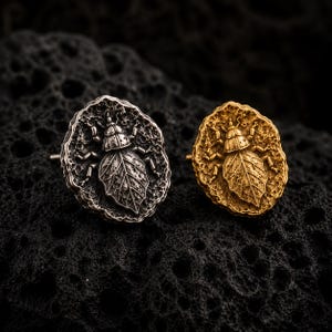 May include: Two stud earrings featuring a detailed insect design with a leaf-shaped body. One earring is finished in a polished silver tone, while the other is finished in a warm gold tone. Both pieces have a textured, organic, and irregular border.