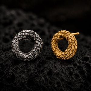 Könnte beinhalten: Zwei kreisförmige Ohrstecker in Form eines gewundenen Drachen oder einer Schlange mit detaillierten Schuppen und einem sichtbaren Kopf. Ein Ohrstecker ist in poliertem Silberton gehalten, der andere in Goldton, beide mit einem Stiftverschluss.