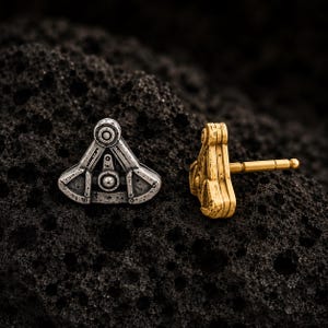 May include: Two stud earrings with a triangular, mechanical-inspired design. One earring is finished in a weathered silver tone, while the other is finished in a gold tone. Each piece features a central circular detail and a post backing.