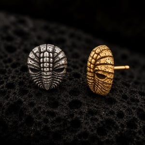 Op de afbeelding: Twee oorknopjes met een getextureerd, gesegmenteerd ontwerp geïnspireerd op trilobieten. De ene oorbel is afgewerkt in een gepolijste zilverkleur, terwijl de andere een goudkleurige afwerking heeft. Elk stuk heeft een ronde, organische vorm.