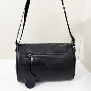 May include: A black cylindrical crossbody bag featuring a textured crocodile-embossed pattern, a horizontal zippered front pocket with a silver-toned pull, and a matching black shoulder strap. A small, scalloped-edge leather tag hangs from the zipper.