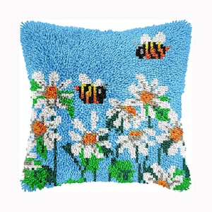 May include: A square throw pillow featuring a textured latch hook design on a light blue background. The pattern displays white daisies with orange centers, green stems and leaves, and two bumblebees with yellow and black striped bodies.