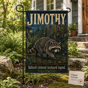 May include: A rectangular garden flag hanging on a black metal stand. The flag features a raccoon in a forest setting with a wooden fence, ferns, and blackberries. The text reads JIMOTHY at the top and Ballard's beloved backyard legend at the bottom.