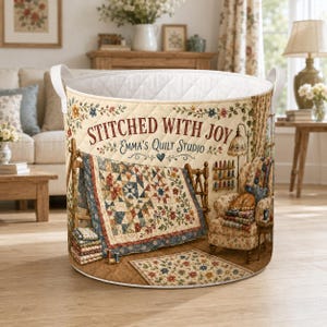 May include: A round, quilted fabric storage basket featuring a printed illustration of a quilt studio with the text STITCHED WITH JOY EMMA'S QUILT STUDIO. The design includes a quilt on a rack, a floral armchair with a cat, and a small rug.