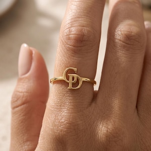 Op de afbeelding: Een goudkleurige ring met een gepolijste afwerking en een centraal ontwerpelement bestaande uit de in elkaar grijpende hoofdletters G en D. De ring is dun en cirkelvormig en heeft een glad, reflecterend oppervlak dat licht vangt.