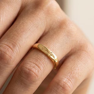 Könnte beinhalten: Ein polierter Goldring mit einer glatten, abgerundeten Oberfläche, in die der Name Emily in einer feinen Schreibschrift eingraviert ist. Der Ring hat eine gleichmäßige Dicke und eine reflektierende Metalloberfläche mit schlichtem Design.