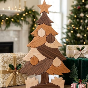 May include: A wooden Christmas tree puzzle featuring alternating light and dark wood grain segments. The tree is topped with a star and decorated with circular ornament shapes. Two small gift-shaped wooden blocks sit at the base of the tree.