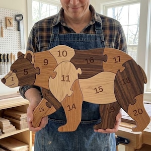 May include: A wooden bear-shaped jigsaw puzzle made of various light and dark wood tones. The puzzle pieces are numbered from 1 to 20, with each piece interlocking to form the complete bear figure. The wood grain is visible on each piece.