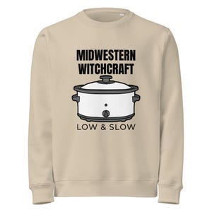 Puede incluir: Una sudadera beige de cuello redondo con un gráfico de una olla de cocción lenta blanca con tapa y base negras. El texto MIDWESTERN WITCHCRAFT está impreso en negro arriba y el texto LOW & SLOW está impreso en negro debajo de la olla.