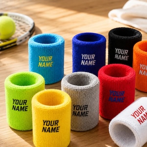 Könnte beinhalten: Eine Sammlung von neun farbenfrohen Frottee-Schweißbändern in Hellblau, Dunkelblau, Schwarz, Orange, Limettengrün, Gelb, Grau, Rot und Weiß. Jedes Schweißband ist mit dem gestickten Text YOUR NAME in einer Kontrastfarbe versehen.
