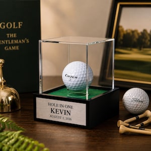 Könnte beinhalten: Ein weißer Golfball mit der Aufschrift Caizer 2 in einer transparenten Acrylvitrine auf einem schwarzen Sockel. Ein silbernes Schild auf der Vorderseite trägt die Aufschrift HOLE IN ONE KEVIN AUGUST 1, 2026. Ein zweiter Golfball und Tees liegen daneben.