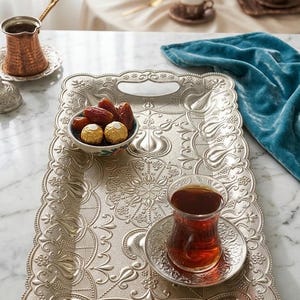 May include: A rectangular silver-toned metal serving tray featuring an intricate embossed floral and geometric pattern with two integrated handles. The tray holds a small bowl of dates and gold-wrapped chocolates, and a glass of tea on a matching saucer.