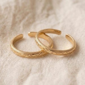 Könnte beinhalten: Zwei goldfarbene offene Ringe mit einem feinen, sich wiederholenden Gravurmuster auf dem äußeren Band. Die Ringe haben eine polierte Oberfläche und ein kreisförmiges, verstellbares Design mit einer kleinen Lücke und filigranen Details.