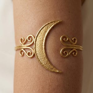 May include: A gold-toned cuff bracelet featuring a central crescent moon shape with a textured surface. The band extends on both sides into symmetrical, open-work scroll designs that terminate in curved, decorative flourishes at the open ends.