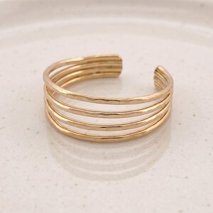 Könnte beinhalten: Ein goldfarbener offener Ring, der aus fünf parallelen Bändern mit einer gehämmerten, texturierten Oberfläche besteht. Der Ring hat eine kreisförmige Form mit einer kleinen Lücke an der Rückseite, was einen gestapelten Look erzeugt. Die Metalloberfläche reflektiert das Licht und betont die unebene, handgefertigte Textur jedes einzelnen Bandes.