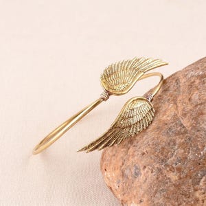 Puede incluir: Una pulsera de metal en tono dorado con dos adornos simétricos en forma de ala en los extremos abiertos. Las alas tienen texturas de plumas grabadas detalladas en sus superficies, y la banda es un alambre fino y liso curvado en forma circular.
