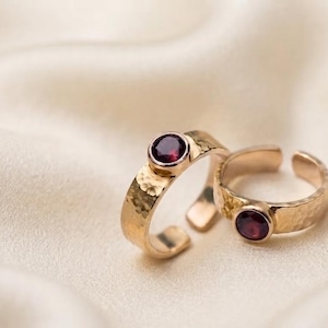 Puede incluir: Dos anillos abiertos de tono dorado con una textura martillada en la banda. Cada anillo tiene una piedra preciosa redonda de color rojo intenso en un engaste de bisel. Los anillos son ajustables, con una abertura en la parte posterior.