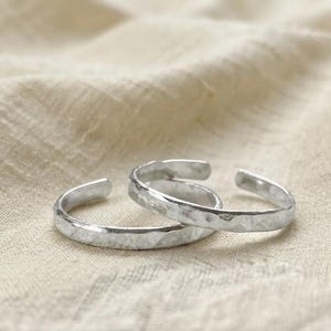 Puede incluir: Dos anillos abiertos de color plateado con una superficie martillada y texturizada. Los anillos tienen un diseño de banda circular con una pequeña abertura, creando una apariencia ajustable. El metal tiene un acabado pulido y reflectante.