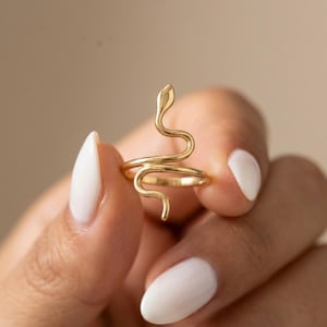May include: A gold-toned ring shaped like a coiled snake. The metal band wraps around the finger, with the snake's head extending upward and the tail curving along the bottom of the band. The surface of the ring is smooth and polished.