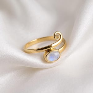 Könnte beinhalten: Ein goldfarbener Bypass-Ring mit einer glatten, polierten Ringschiene, die an einem Ende in einer Spirale endet und am anderen Ende einen ovalen, in einer Zargenfassung gefassten, irisierenden Mondstein-Cabochon mit bläulichem Schimmer trägt.