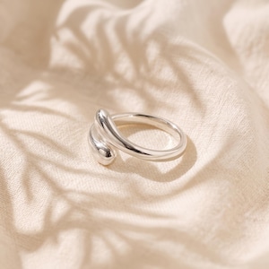 Op de afbeelding: Een ring van gepolijst zilver met een open bypass-ontwerp met twee afgeronde, taps toelopende uiteinden die elkaar licht overlappen. Het metalen oppervlak is glad en reflecterend en vormt een doorlopende, vloeiende vorm rond de vinger.