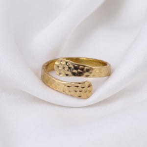 Puede incluir: Un anillo abierto de tono dorado con una textura martillada en toda la superficie. La banda tiene dos extremos redondeados y superpuestos que crean un diseño cruzado, mostrando un acabado metálico reflectante con hendiduras.