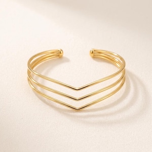 Puede incluir: Un brazalete tipo puño de tono dorado con un diseño de chevrón de tres capas. La pulsera abierta está construida con tres bandas metálicas delgadas, lisas y paralelas que se encuentran en un punto central, creando una silueta en forma de V.