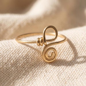 Könnte beinhalten: Ein goldfarbener Drahtring mit einem zentralen Design, das aus einer kleinen kreisförmigen Spirale und einem größeren geschlungenen Drahtelement besteht. Der Ring ist aus einem einzigen Stück dünnem Metalldraht gefertigt, der sich selbst umschlingt.