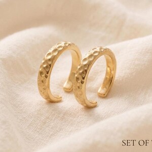 Pode incluir: Um conjunto de dois brincos ear cuff dourados com uma superfície martelada e texturizada. Cada peça tem um design de banda aberta com acabamento polido. O texto "SET OF TWO" está impresso no canto inferior direito da imagem.