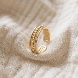 May include: A gold-toned open ring featuring a central band of raised spherical beads flanked by two thin twisted rope-style borders. The metal surface has a polished finish, and the band is adjustable with a gap at the back of the ring.