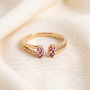May include: A gold open-style ring featuring two split ends, each set with two small, round, vibrant pink gemstones. The band has a smooth, polished finish, and the four stones are arranged in pairs at the opening of the ring's circular band.