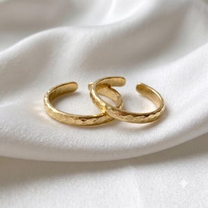 Könnte beinhalten: Zwei goldfarbene offene Ringe mit einer texturierten, gehämmerten Oberfläche. Die Ringe haben ein kreisförmiges Banddesign mit einer kleinen Lücke, was eine verstellbare Größe ermöglicht. Das Metall hat eine reflektierende, polierte Oberfläche.