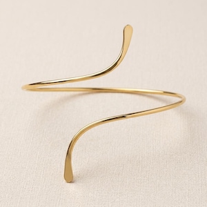 May include: A gold-toned arm cuff featuring a sleek, open-ended spiral design. The metal band has a smooth, polished finish and curves into two tapered ends that extend in opposite directions, creating a continuous, fluid, and minimalist shape.