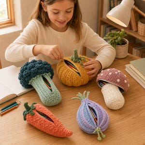 Può includere: Una collezione di cinque astucci all'uncinetto a forma di cibo, tra cui un broccolo blu scuro, una zucca gialla, un fungo rosa con pois bianchi, una carota arancione e una cipolla viola chiaro, ognuno dotato di un'apertura con cerniera.