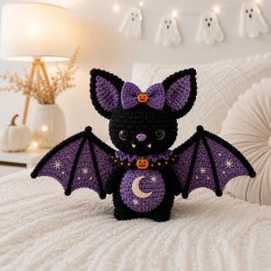 May include: A black and purple crocheted bat plush toy featuring large ears with a purple bow, a small orange pumpkin accent, a ruffled collar, and wings decorated with white star embroidery. The belly has a purple patch with a crescent moon.