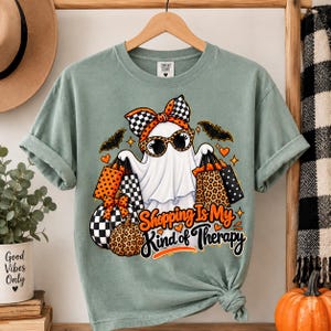 Puede incluir: Una camiseta verde salvia con un gráfico de un fantasma con un lazo a cuadros y gafas de sol de leopardo, sosteniendo bolsas de compras. El diseño incluye calabazas y el texto Shopping Is My Kind of Therapy en letras naranjas y negras.