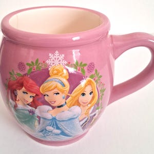 Puede incluir: Taza de cer&aacute;mica rosa con tres princesas de dibujos animados. La taza tiene forma redondeada con asa e interior rosa claro. Las princesas est&aacute;n representadas en un dise&ntilde;o colorido con tem&aacute;tica invernal.